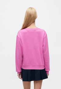 Ροζ φούτερ με στρογγυλή λαιμόκοψη, χαμηλωμένους ώμους και ριμπ μανσέτες, συνδυασμένο με μπλε σκούρα πλισέ φούστα. Ομαλή υφή βαμβακιού.