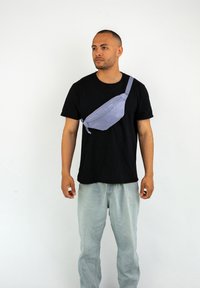Sac bandoulière violet clair en tissu, doté d'une fermeture éclair et d'une sangle réglable, porté sur un t-shirt noir et un jean clair.