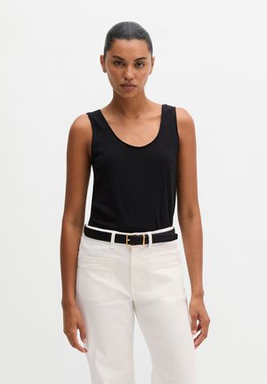 Frau mit dunklem Haar in einem schwarzen ärmellosen Oberteil und weißen Hosen, stehend vor einem schlichten weißen Hintergrund.