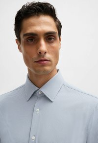 Chemise boutonnée rayée bleu clair et blanche avec un col ouvert, présentant un motif subtil et une texture de tissu lisse.