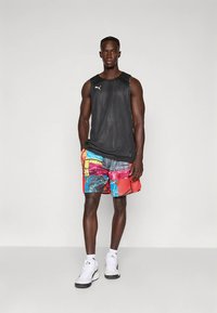 Vīrietis, ģērbies melnā bezroku Puma krekliņā, krāsainās grafisko motīvu šortos, baltās Puma zeķēs un baltos sporta apavos, stāvot pret vienkāršu pelēku fonu.