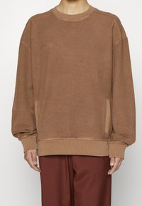 Sudadera marrón con cuello redondo, puños acanalados y bolsillos laterales. Presenta el logotipo "HUGO" en tono y un acabado texturizado. Diseño de corte holgado.