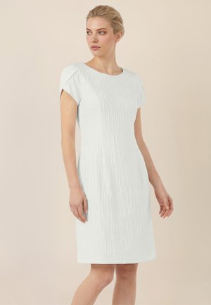 Young woman stands wearing a white, short-sleeved, textured knee-length dress against a plain beige background.