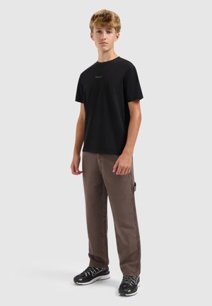 Teen wearing a black short-sleeve t-shirt, brown loose pants with a side loop, and black athletic shoes on a plain background.