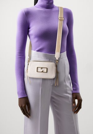 Person iført en tætsiddende lilla rullekrave og lysegrå højttaljede bukser med en cremefarvet crossbody-taske med guldfarvet hardware.