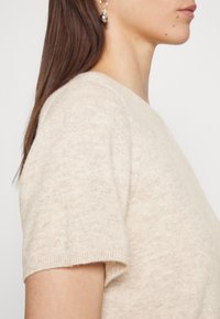 Beige kortærmet sweater i blød, tekstureret strik. Rund halsudskæring og ribbede manchetter. Glat stof med minimale variationer.