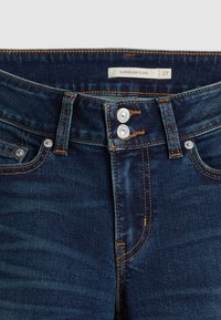 Jean en denim bleu foncé avec double boutons argentés, coutures orange, poches avant, passants de ceinture et une étiquette de taille indiquant "SUPERLOW FLARE 27".