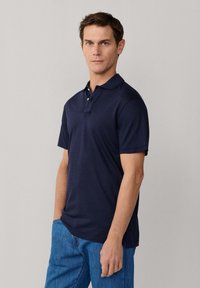 Hombre con camisa polo azul marino y jeans azules, de pie con una mano en el bolsillo contra un fondo claro y liso.