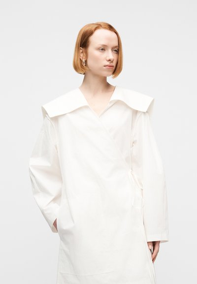 Γυναίκα με κοντά κόκκινα μαλλιά που φοράει ένα λευκό, φαρδύ φόρεμα με φαρδιά γιακά και μακριά μανίκια, στέκεται μπροστά σε μια απλή φόντο.