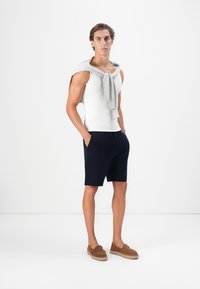 Mann steht mit den Händen in den Taschen, trägt ein weißes Tanktop, marineblaue Shorts, einen grauen Pullover über den Schultern und braune Slipper.