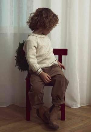 Adīta krēmkrāsas džemperis ar plankumiem, brūnas korduroja bikses un brūni potīšu zābaki, sēžot uz sarkanas krēsla pret gaismas aizkariem.