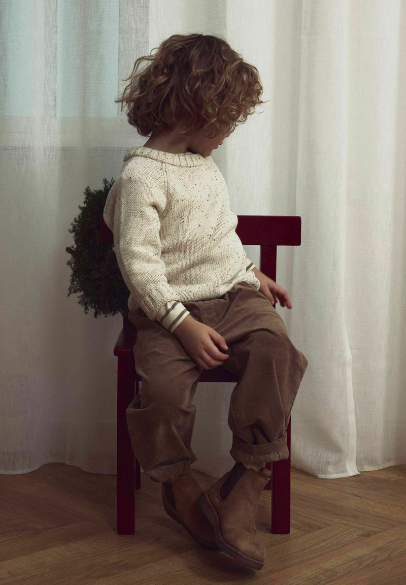 Pleten pulover v kremni barvi s pikicami, rjave kordflavne hlače in rjavi gležnjarji, sedeč na rdečem stolu ob svetlih zavesah.