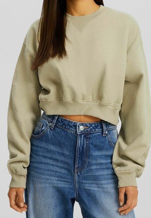 Personne portant un sweat-shirt court beige à manches longues et un jean denim bleu taille haute, les mains relâchées sur les côtés.