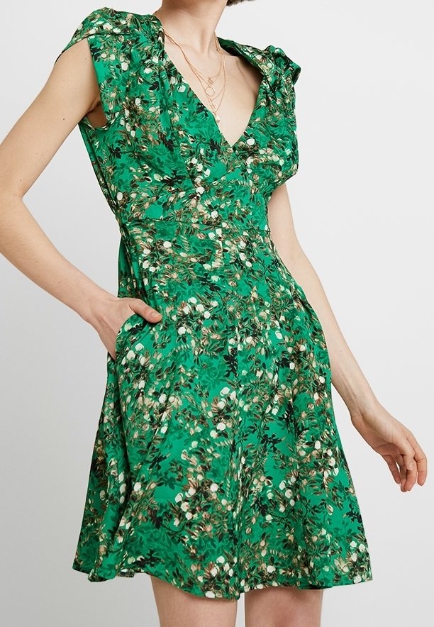 Robe verte à fleurs avec col en V, manches courtes et taille cintrée, portée avec des colliers dorés superposés, main dans la poche d'un côté.