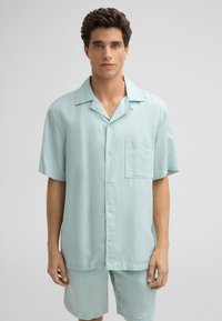 Helles blaues Kurzarmhemd aus einem glatten, leichten Stoff, mit einer einzelnen Brusttasche und durchgehender Knopfleiste.