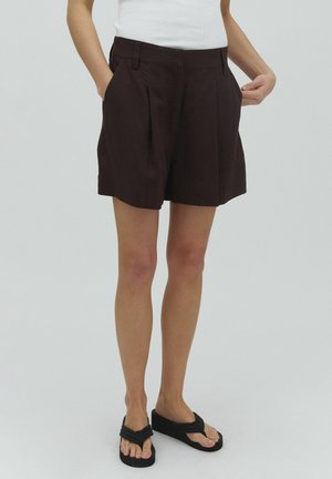 Vrouw draagt donkerbruine geplooide korte broek, witte mouwloze top en zwarte platformslippers, staand met handen in de zakken tegen een effen achtergrond.