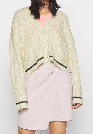 Kobieta ubrana w pastelowożółty krótki kardigan w czarne paski, na różowym topie i jasnoróżowej owijanej spódnicy, na jednolitym tle.