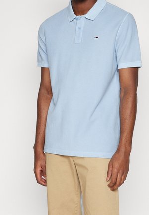 Mann trägt hellblaues kurzärmeliges Poloshirt mit kleinem Logo auf der Brust und beigefarbene Hose, steht vor einfachem Hintergrund.