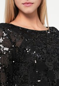 Top preto com lantejoulas, apresentando uma textura suave, um corte descontraído e um decote largo. As lantejoulas criam um detalhe cintilante e refletor.