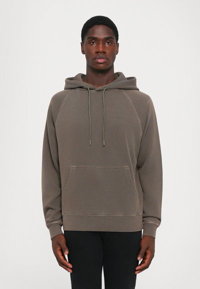 Junger Mann in einem grauen Kapuzenpullover mit vorderer Tasche und schwarzen Hosen, der vor einem schlichten weißen Hintergrund steht.