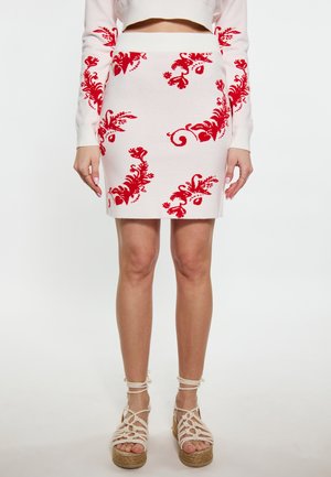 Kvinde iført en hvid nederdel med røde blomsterprint kombineret med en hvid langærmet top og beige kilehælssandaler med snører af reb.