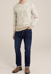 Man i beige blommönstrad tröja, mörkblå jeans med uppvikta ben och bruna mockastövlar, står med en hand i fickan.