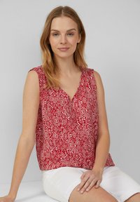 Rode mouwloze top met een geometrisch wit patroon en een V-hals, gemaakt van lichtgewicht stof. Draagt met witte shorts.