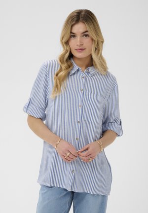 Kobieta o długich falowanych blond włosach, ubrana w niebiesko-białą, paskowaną koszulę zapinaną na guziki z podwiniętymi rękawami oraz jasnoniebieskie dżinsy.