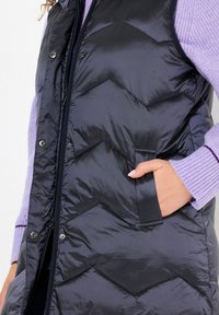 Fyllig mörkblå väst med chevronstygning, dragkedja och tryckknappar. Har sidofickor och är lagd över en ljuslila tröja.