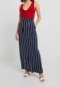 Rode mouwloze top gecombineerd met een marineblauwe maxi rok met witte verticale strepen en zijzakken, aangevuld met witte sandalen.