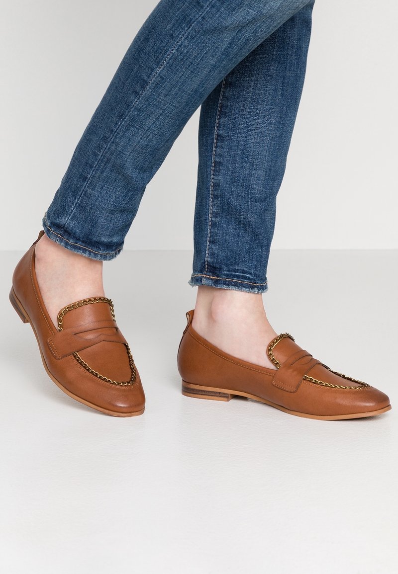 Pieds portant des mocassins en cuir marron clair avec détail chaînette dorée, associés à un jean bleu sur un fond uni.
