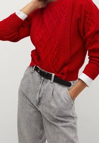 Roter Kabelstrickpullover kombiniert mit hoch geschnittenen grauen Jeans. Das Outfit wird durch einen schwarzen Gürtel mit einer silbernen Schnalle ergänzt.
