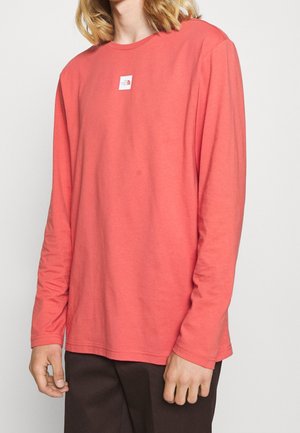 T-shirt corail à manches longues en tissu doux, avec un col rond et une petite étiquette de logo sur la poitrine. Design simple et droit.