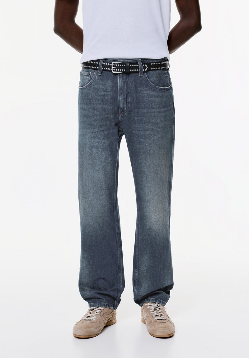 Ljusblå denimjeans med rak passform, prydda med ett svart nitarbälte. Beige sneakers fulländar outfiten, mot en vit bakgrund.