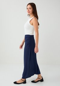 Marineblaue lange Rock mit gerafftem Taillendetail, kombiniert mit einem weißen, körperbetonten Tanktop und schwarzen Ballerinas. Glatter, leichtes Material.