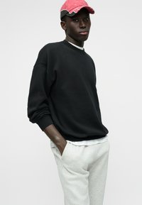 Sudadera negra con hombros caídos, combinada con pantalones de chándal gris claro. Una gorra rosa con visera curva completa el conjunto.