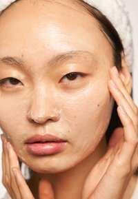 Masque de soin facial appliqué sur le visage ; texture lisse et crémeuse ; couleur pâle ; modèle touchant son visage avec la main, maquillage neutre.