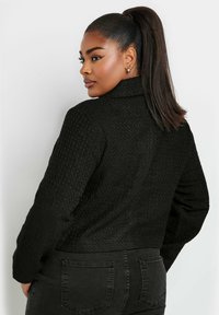 Svart texturerad jacka med krage, med långa ärmar och en kortad passform. Utsmyckad med ett subtilt mönster och öppen ryggdesign.