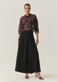 Zwarte wijde broek gecombineerd met een doorschijnende blouse met een donkere bloemenprint, voorzien van een kraag en knoopsluiting. Eenvoudig, elegant ontwerp.
