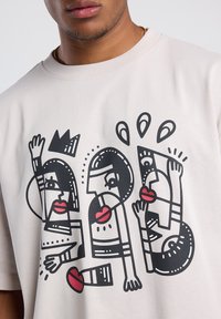 Light beige cotton t-shirt featuring a large black graphic print of abstract faces with red lips and geometric accents. Crew neckline.