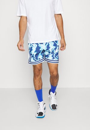 Person trägt hellblaue und weiße Basketballshorts mit Blitzmuster, weißes Shirt, blaue Sportsocken und weiße Sportschuhe.