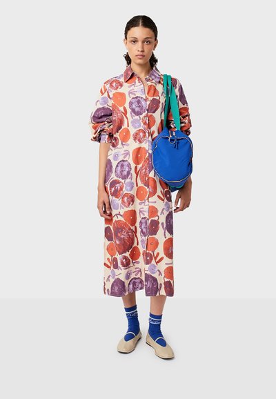 Młoda kobieta w długiej kwiecistej sukience z podwiniętymi rękawami, w niebieskich skarpetach, w beżowych butach, niosąca jasnoniebieską torbę z zielonym paskiem.