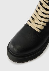 Črne usnjene čevlje z teksturiranim zaključkom, okroglo konico in bež vezalkami. Podplat je debele in črn ter ima vzorčasto podlago za boljši oprijem.