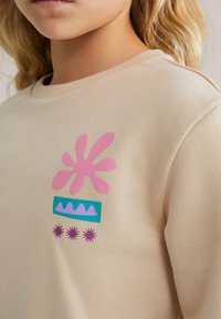 Tuvo plāna beige krēklā ar abstraktiem rozā, zaļiem un purpursarkaniem motīviem uz krūtīm, ko valkā blondi mati cilvēks.