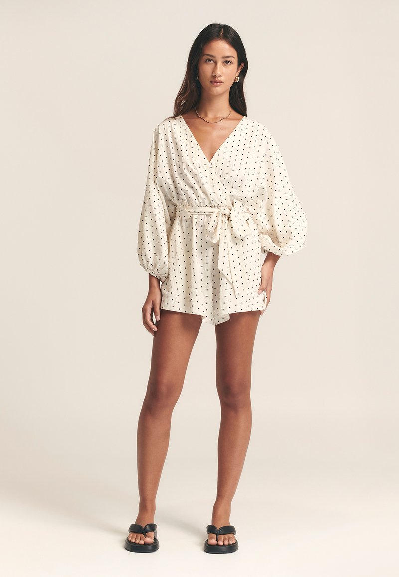 Romper blanc à pois avec un design croisé, des manches bouffantes et une taille nouée. Présente un décolleté en V et un design de short ajusté.