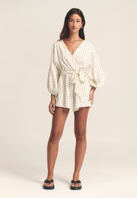 Hvidt prikket slå-om-playsuit med lange ballonærmer, bindebånd i taljen og shorts. Modellen har sorte sandaler på.