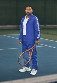 Kék sportos tréningruha fehér kiegészítőkkel, amely zipzáros dzsekiből és elasztikus derekú nadrágból áll. Egy narancssárga teniszütőt és egy zöld teniszlabdát tart.