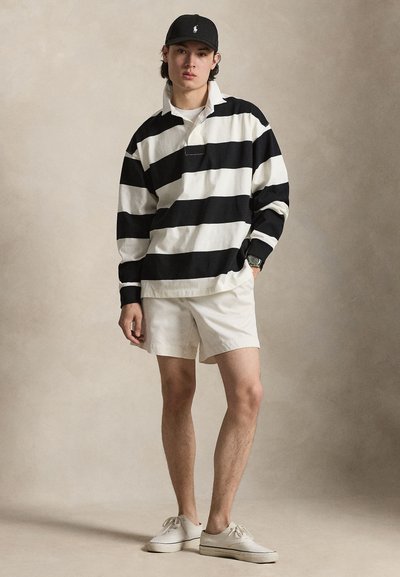 Jonge man die staat en een zwart-wit gestreept shirt met lange mouwen, witte korte broek, witte sneakers en een zwarte pet draagt.