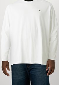 Man med en vit skjorta med långa ärmar och en liten grön krokodil-logotyp på vänster bröst, samt mörkblå jeans, stående mot en vit bakgrund.