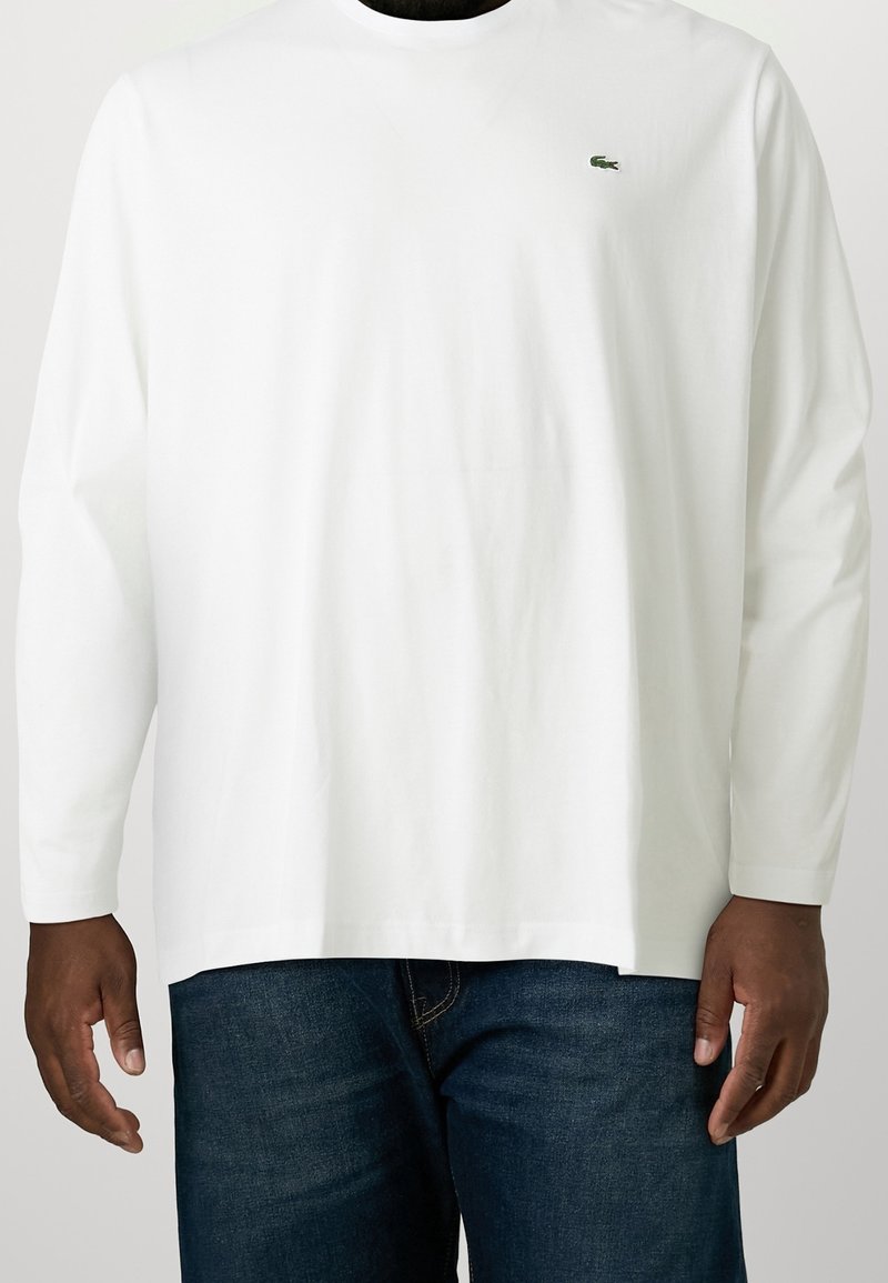 Man med en vit skjorta med långa ärmar och en liten grön krokodil-logotyp på vänster bröst, samt mörkblå jeans, stående mot en vit bakgrund.